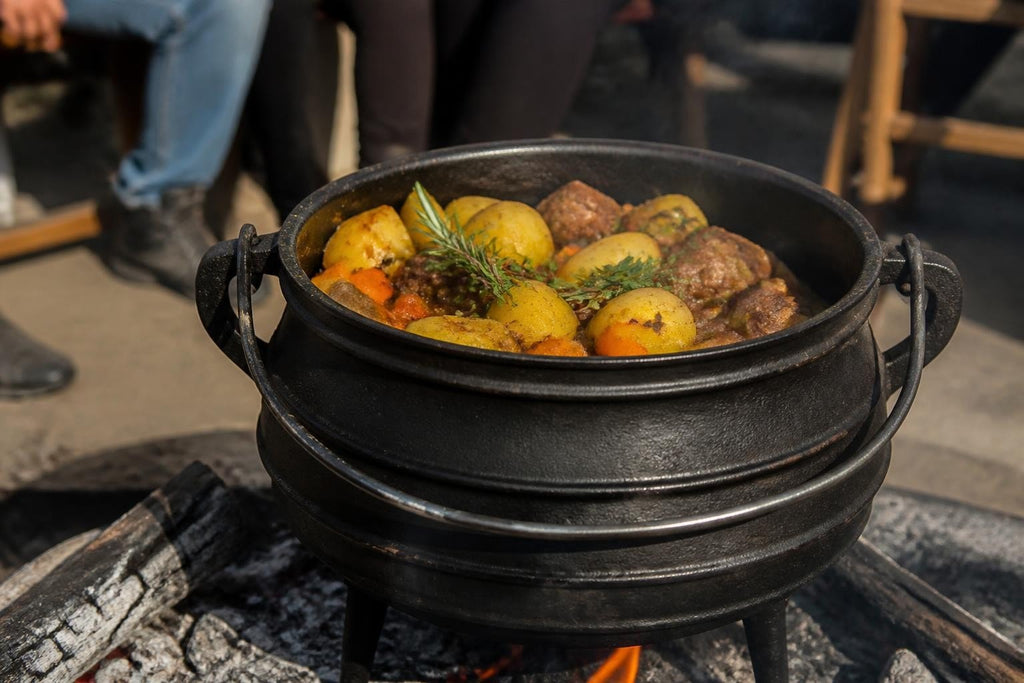 Cast-iron potjie with a black three-legged pot cooking meat, potatoes and herbs, Potjie: South African Slow-Cooked Tradition