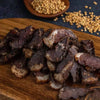 Sliced original South African biltong on wooden board with coriander seeds in background, ready-to-eat air-cured dried meat snack.