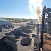 Industrial charcoal production site with large piles of black charcoal and machinery emitting smoke under a clear sky