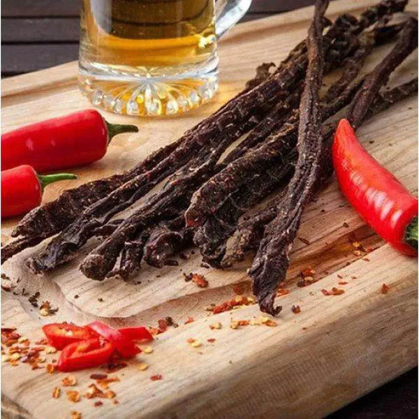 Spicy ready-to-eat biltong chilli sticks on wooden board with red chili peppers and beer glass