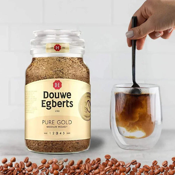 Douwe Egberts Pure Gold 200g instant coffee jar with medium roast label next to a glass cup being stirred and coffee beans scattered on surface