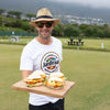 Man outdoors holding wooden board with grilled chicken sandwiches, promoting Jan Braai Prego Sauce for basting and marinades