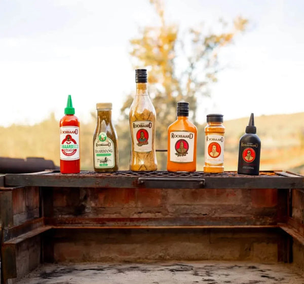 Rooibaard Kwaadbaard Sauce bottles displayed outdoors on a rustic surface.