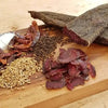 Slices of original South African biltong on a wooden board with dried chili peppers, black peppercorns, and coriander seeds