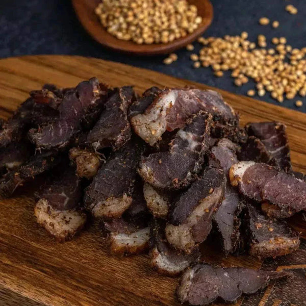Sliced original South African biltong on wooden board with coriander seeds in background, ready-to-eat air-cured dried meat snack.