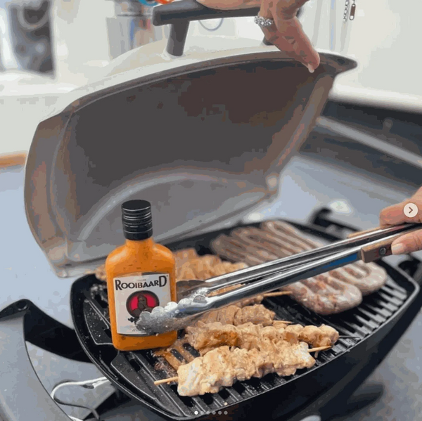Rooibaard Original Sauce 250ml bottle next to grilling meat on barbecue.