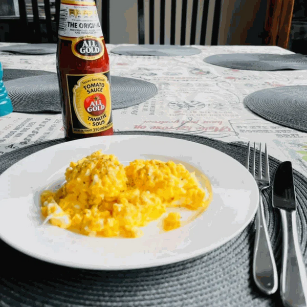 All Gold Tomato Sauce 700ml bottle next to a plate of scrambled eggs on a set dining table.