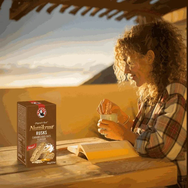 Nuttikrust Caramelised Oats Rusks 450g box next to woman dipping rusk in cup by open book at sunrise