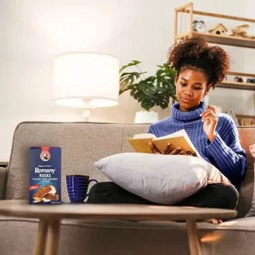 Woman enjoying a cozy evening with a book and Bakers Romany Classic Choc Coconut Flavour Rusks on the table.