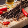 Spicy ready-to-eat biltong chilli sticks on wooden board with red chili peppers and beer glass