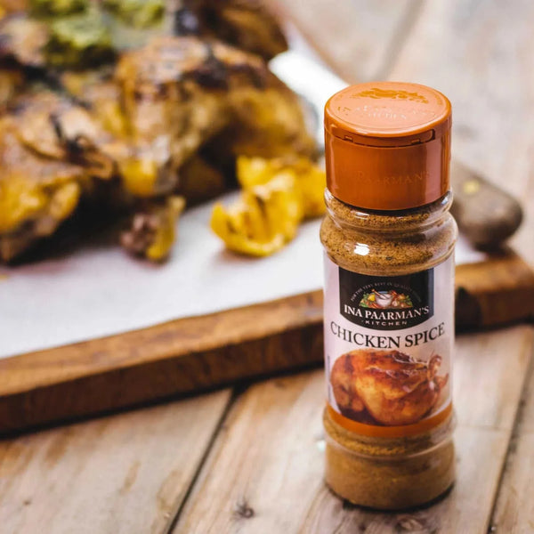 Ina Paarman Chicken Spice bottle on wooden table with roasted chicken in background.