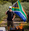Jan Braai holding South African flag next to a barbecue fire in an outdoor setting.