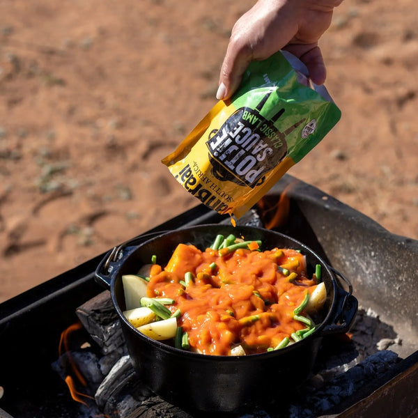 Jan Braai Sauces Classic Lamb Potjie Sauce being poured over vegetables in potjie pot over coals outdoors